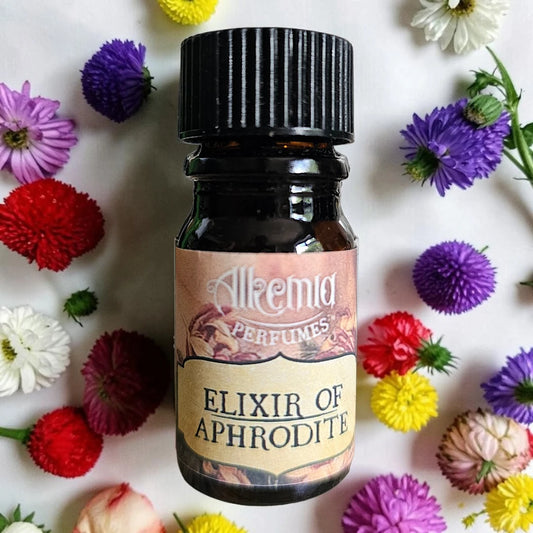 A small amber glass bottle of Alkemia Perfumes 'Elixir of Aphrodite' with a black ribbed cap and a beige label, centered on a white background and surrounded by colorful artificial flowers.
Alkemia Elixir Of Aphrodite fragrance samples UK 1ml sample available 