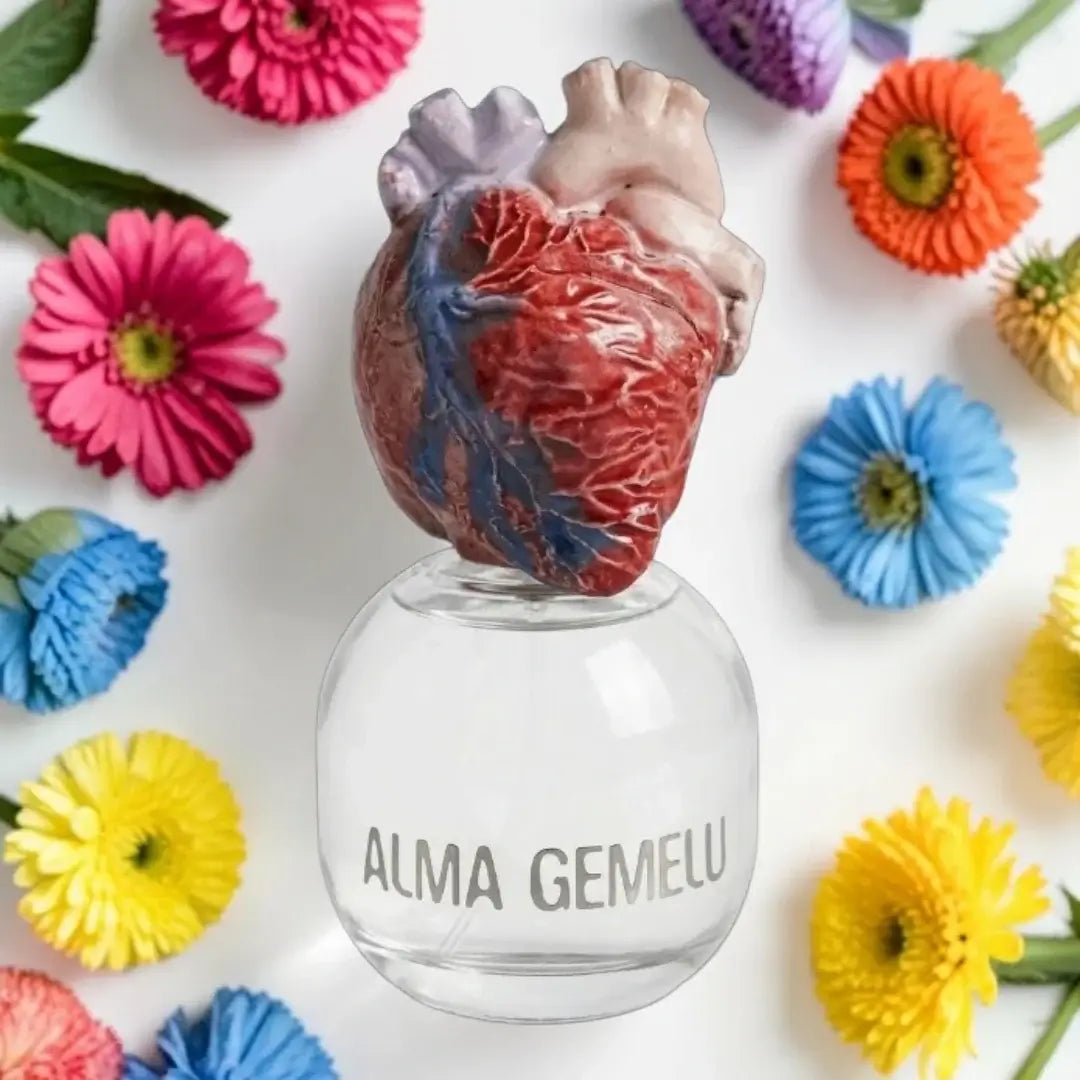 A clear glass bottle with the words 'ALMA GEMELU' printed on the front, topped with a realistic anatomical heart sculpture. The bottle is photographed from above on a white background, surrounded by colorful artificial flowers.