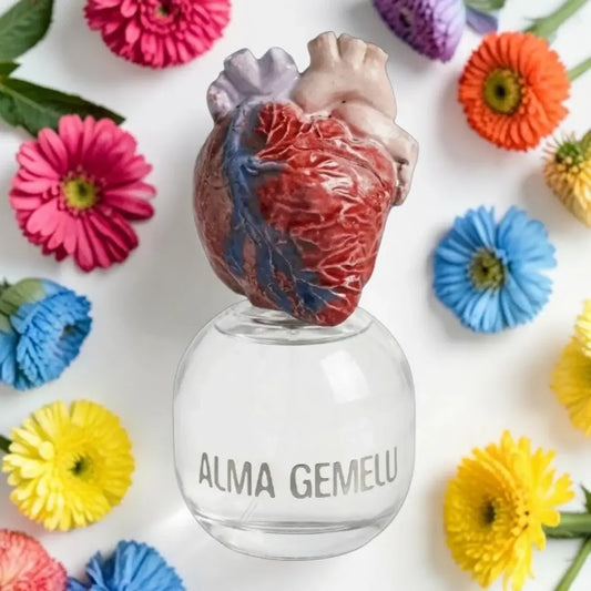 A clear glass bottle with the words 'ALMA GEMELU' printed on the front, topped with a realistic anatomical heart sculpture. The bottle is photographed from above on a white background, surrounded by colorful artificial flowers.