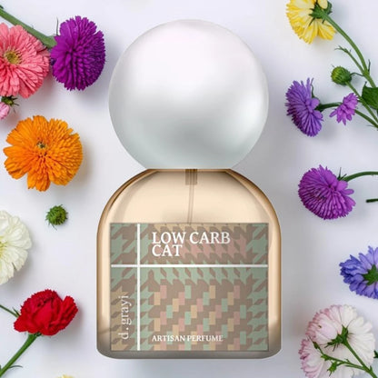 A perfume bottle labeled 'LOW CARB CAT' by d'Grayv, styled in a flat-lay with colorful flowers around it on a white background.