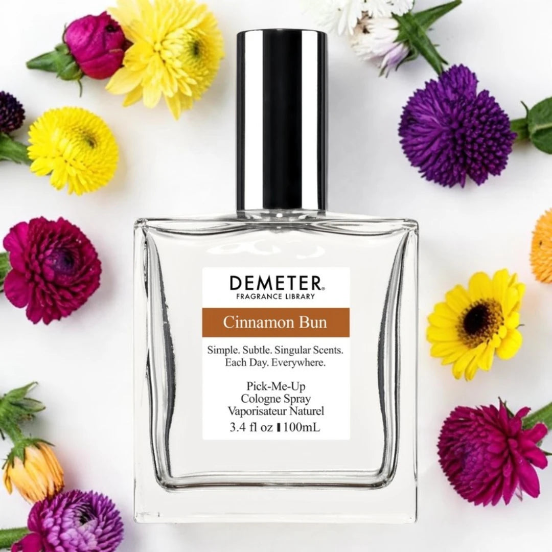 A bottle of Demeter Fragrance Library Cinnamon Bun cologne spray, surrounded by colorful flowers on a white background.