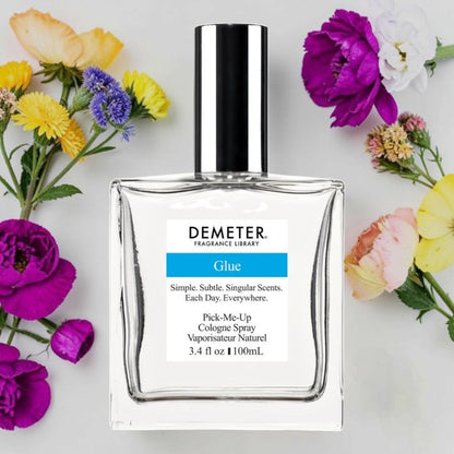 A bottle of Demeter Fragrance Library Glue cologne spray, surrounded by colorful flowers on a light gray background. Demeter Fragrance Library Glue | Fragrance Samples UK 1ml 2ml and 5ml perfume samples available 
