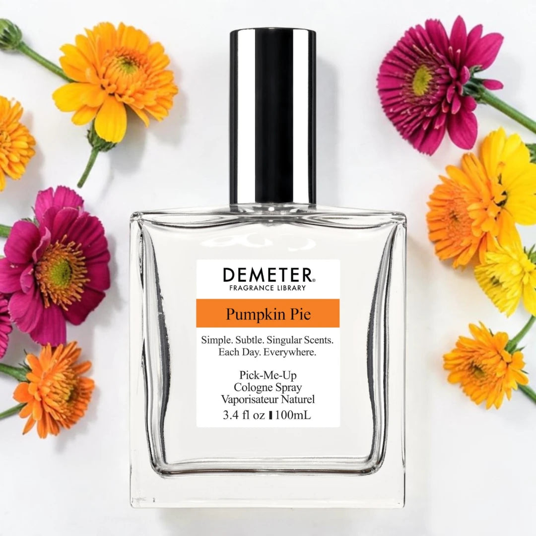 A bottle of Demeter Fragrance Library 'Pumpkin Pie' cologne spray, styled in a flat-lay with colorful orange, yellow, and pink flowers on a white background.