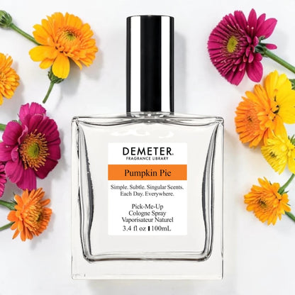A bottle of Demeter Fragrance Library 'Pumpkin Pie' cologne spray, styled in a flat-lay with colorful orange, yellow, and pink flowers on a white background.