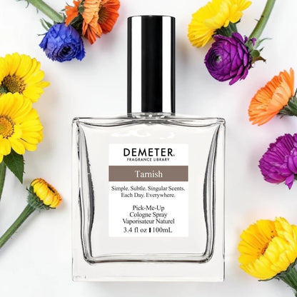 A bottle of Demeter Fragrance Library 'Tarnish' Cologne Spray, presented in a clear glass bottle with a black cap, surrounded by colorful flowers on a white background. Demeter Fragrance Library Tarnish | Fragrance Samples UK