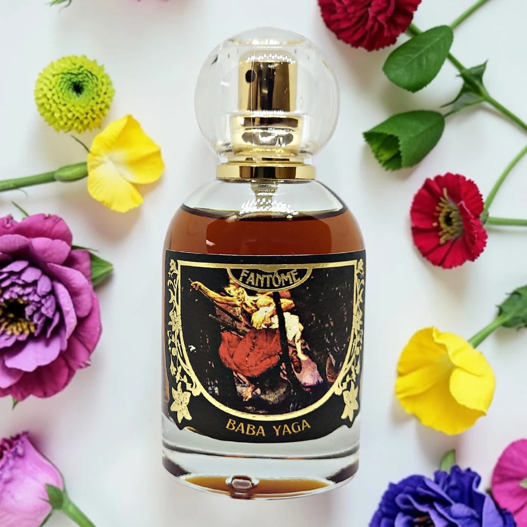 A glass perfume bottle with a clear spherical cap and gold spray mechanism, filled with amber liquid. The bottle has a black label with gold detailing and an illustration, labeled 'FANTOME' and 'BABA YAGA'. available