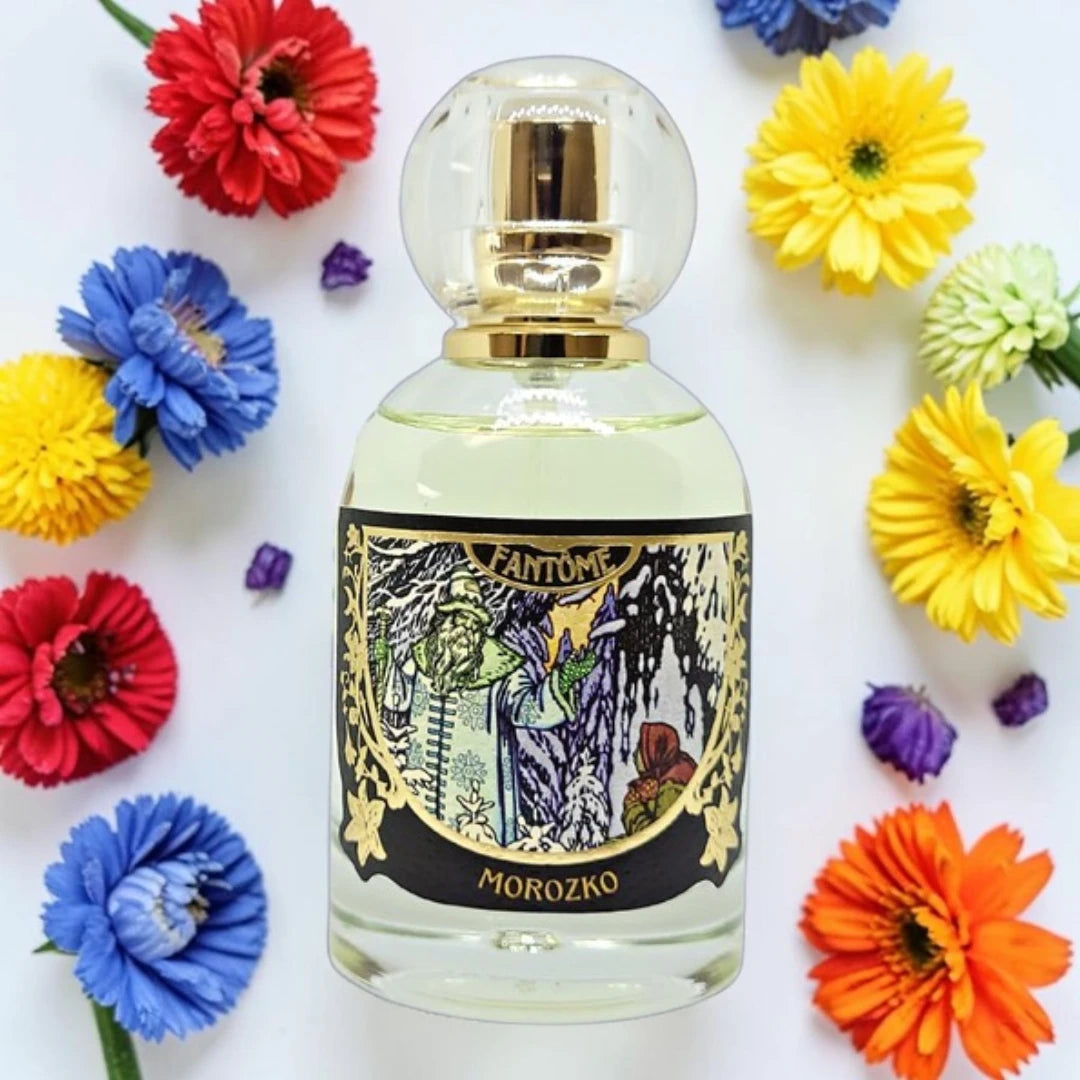 A clear glass perfume bottle with a gold cap and a decorative black label reading 'Fantome' and 'Morozko', surrounded by colorful artificial flowers on a white background. Fantôme Morozko | Fragrance Samples UK 1ml 2ml and 5ml perfume samples available 