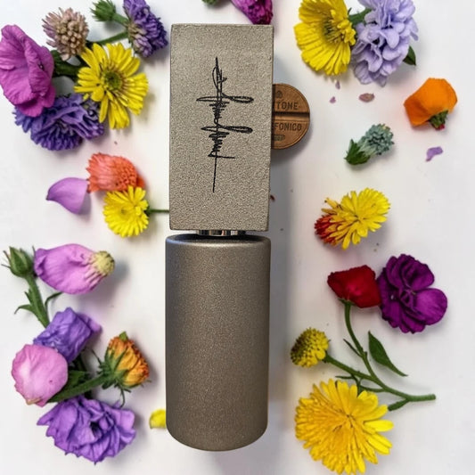 A cylindrical metallic cosmetic tube with a rectangular cap featuring a black abstract symbol, arranged on a white background and surrounded by colorful flowers. Filippo Sorcinelli Gettone | Fragrance Samples UK
