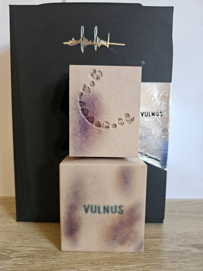 A VULNUS perfume bottle and its packaging box, featuring a beige body with a purple smoky gradient and a metallic gold logo, set against a black box on a wooden surface.
Filippo Sorcinelli Vulnus | Fragrance Samples UK 1ml 2ml and 5ml perfume samples available 