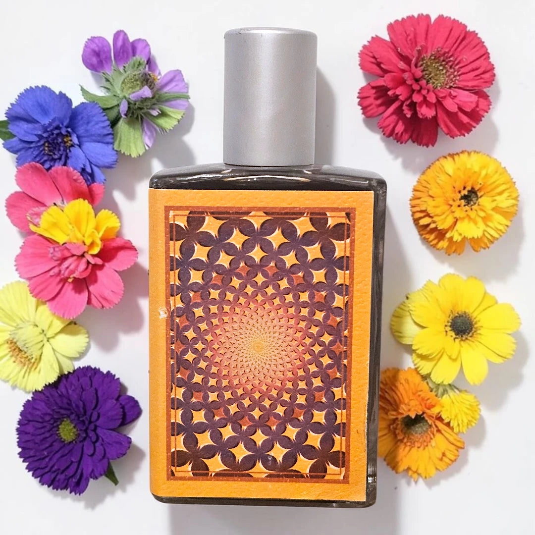 A rectangular perfume bottle with a silver cap and a decorative orange and brown geometric pattern on the front, surrounded by colorful artificial flowers on a white background.
