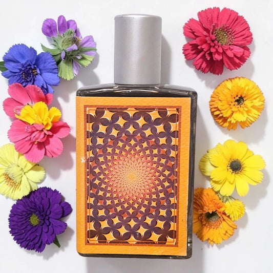 A rectangular perfume bottle with a silver cap and a decorative orange and brown geometric pattern on the front, surrounded by colorful artificial flowers on a white background.