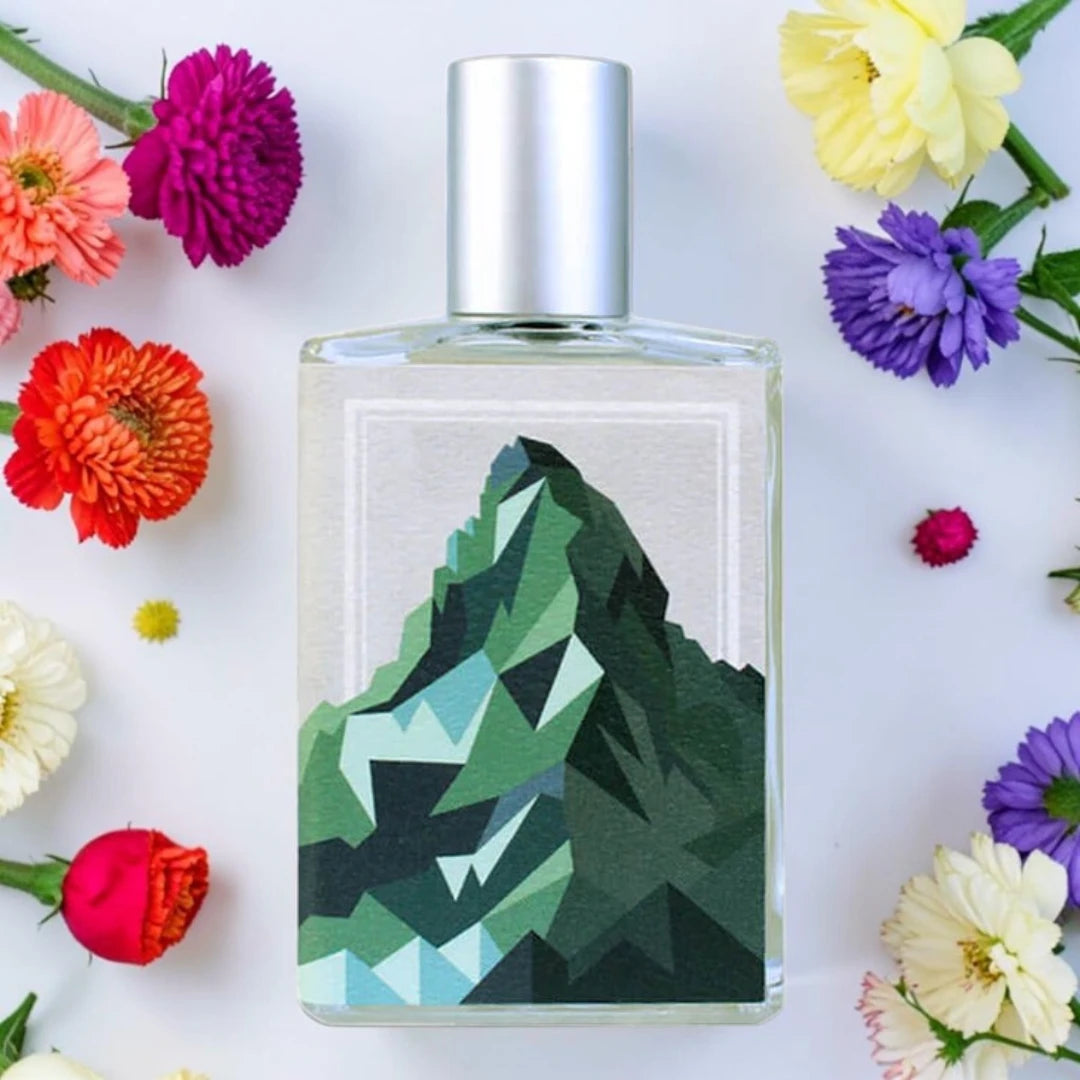 A rectangular glass perfume bottle with a silver cap, featuring a geometric mountain illustration in shades of green and blue on the front label. The bottle is centered on a white background and surrounded by colorful flowers. Imaginary Authors The Language Of Glaciers | Fragrance Samples UK 1ml 2ml and 5ml perfume samples available 