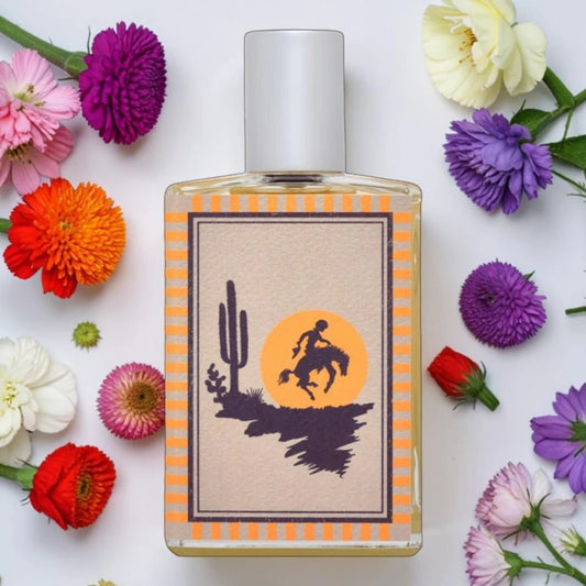 A rectangular glass perfume bottle with a white cap, featuring a desert-themed label with a cactus, silhouetted rider, and orange sun. The bottle is surrounded by colorful flowers on a white background. Imaginary Authors Untamable | Fragrance Samples UK 1ml 2ml and 5ml perfume samples available 