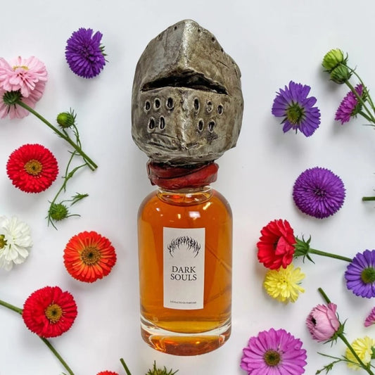 A glass bottle filled with amber liquid, topped with a sculpted medieval knight helmet cap, arranged on a white background and surrounded by colorful daisy flowers. The bottle label reads 'Dark Souls'. Malasombra by Alejando Acosta Dark Souls | Fragrance Samples UK 1ml 2ml 5ml perfume samples