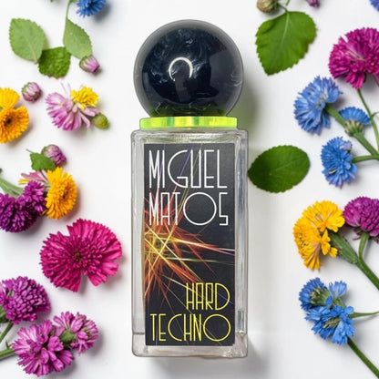 A rectangular glass perfume bottle labeled 'MIGUEL MATOS HARD TECHNO' sits centered on a white background, surrounded by colorful flowers and green leaves. The bottle has a black label with abstract orange and yellow light streaks, a black spherical cap, and a bright yellow-green collar. Fragrance Sample (1ml / 2ml / 5ml)
100% genuine fragrance decanted into a UV-sterilised glass atomiser.
