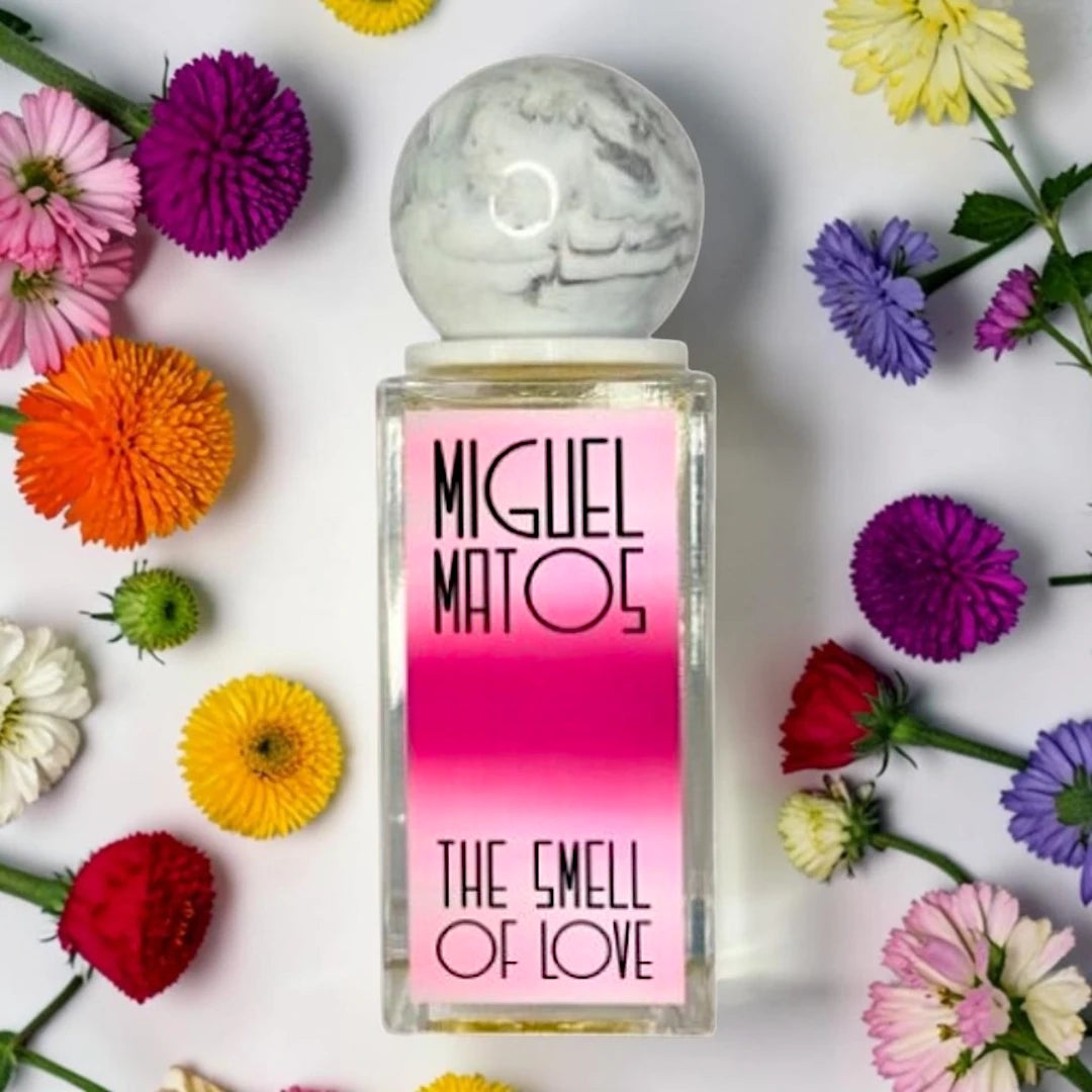 A rectangular glass perfume bottle with a pink-to-magenta gradient label reading 'MIGUEL MATOS' and 'THE SMELL OF LOVE', topped with a round white-and-gray marble cap. The bottle is centered on a white background and surrounded by colorful flowers. Fragrance Sample (1ml / 2ml / 5ml)
100% genuine fragrance decanted into a UV-sterilised glass atomiser.