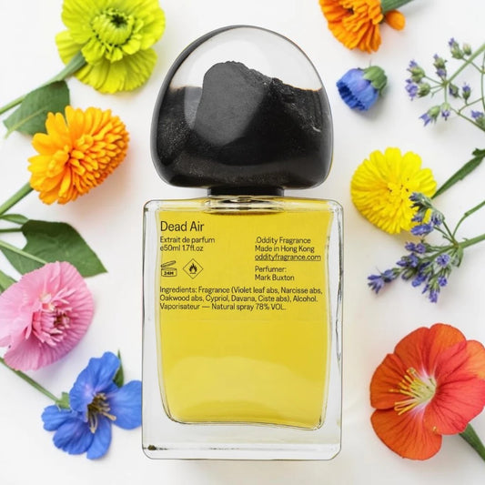 A rectangular glass bottle of Dead Air fragrance with a black dome-shaped cap, filled with yellow liquid, surrounded by colorful flowers on a white background.