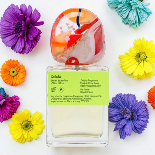 A Delulu perfume bottle with a colorful, fish-shaped cap, surrounded by vibrant artificial flowers on a white background. The bottle has a bright lime green label with black text detailing the fragrance ingredients and creator David Chiche.