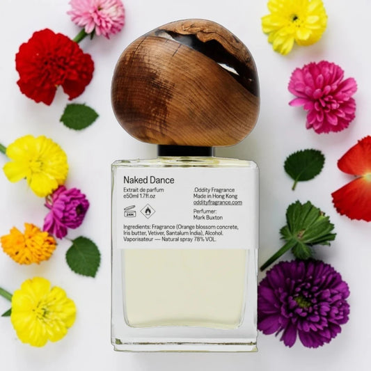 A clear glass bottle of Naked Dance fragrance with a rounded wooden cap, surrounded by colorful flowers on a white background. The label reads 'Naked Dance' and includes fragrance details.