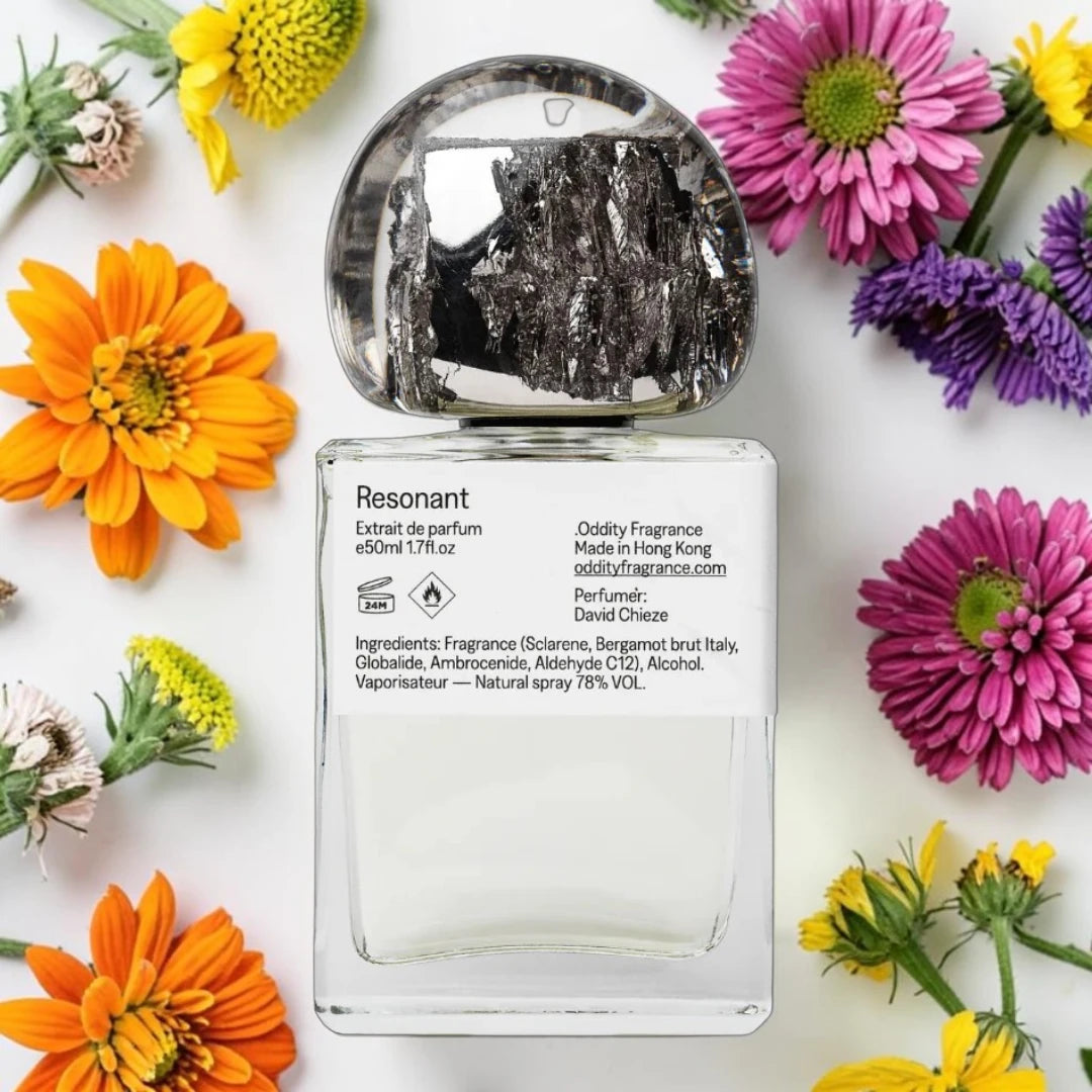 A clear rectangular perfume bottle with a silver cap, labeled 'Resonant' by Oddity Fragrance, styled in a flat lay with colorful flowers on a white background.