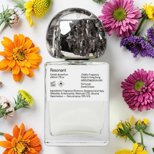 A clear rectangular perfume bottle with a silver cap, labeled 'Resonant' by Oddity Fragrance, styled in a flat lay with colorful flowers on a white background.