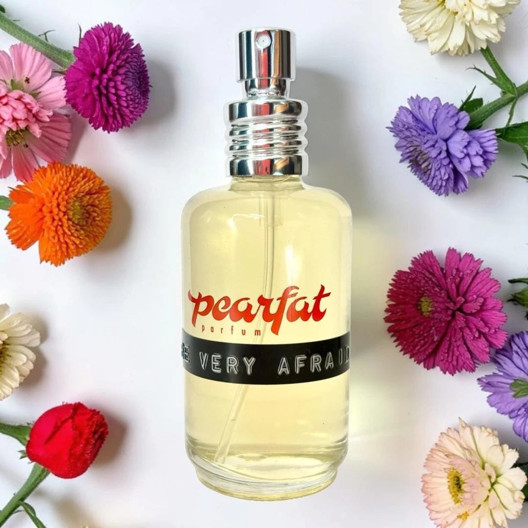 A bottle of Pearfat Perfume with a silver atomizer and a black label reading ' BEVERY AFRAID' is centered on a white background, surrounded by colorful flowers including pink, purple, orange, red, and white. Pearfat Parfum Be Very Afraid | Fragrance Samples UK 1ml 2ml and 5ml perfume samples 