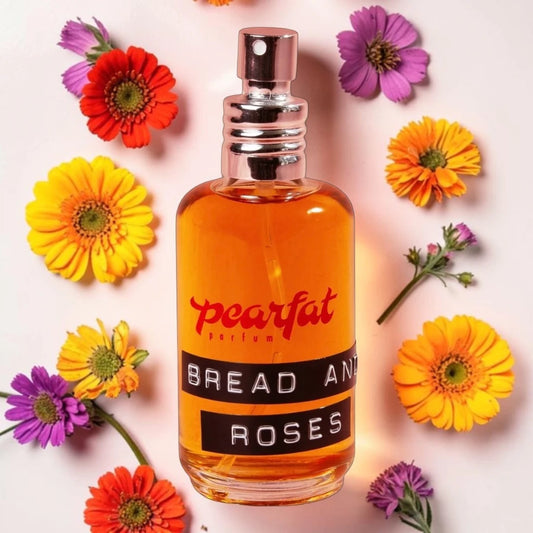 A perfume bottle labeled 'pearlfat parfum BREAD AND ROSES' filled with amber liquid, surrounded by colorful gerbera daisies on a light pink background.