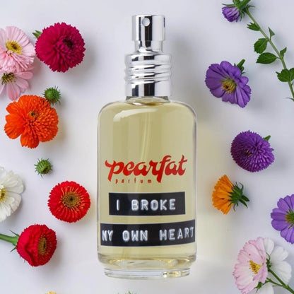 A clear glass perfume bottle with pale yellow liquid and a silver spray top, labeled 'pearlfat perfume I BROKE MY OWN HEART' in red and black text. The bottle is centered on a white background and surrounded by colorful flowers including pink, red, orange, purple, and white blossoms. Pearfat Parfum I Broke My Own Heart | Fragrance Samples UK 1ml 2ml and 5ml perfume samples available 