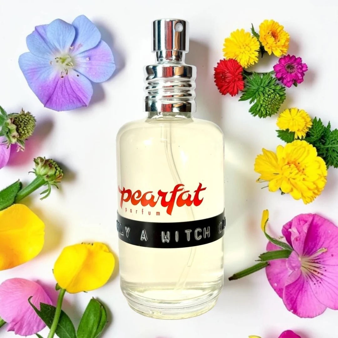 A clear glass perfume bottle with pale yellow liquid and a silver spray top, labeled 'pearfat' in red script with a black band reading 'ONLY A WITCH CAT'. The bottle is centered on a white background and surrounded by colorful flowers including purple, yellow, pink, and red blossoms with green leaves. Pearfat Parfum Only A Witch Cat | Fragrance Samples UK 1ml 2ml and 5ml perfume samples available 