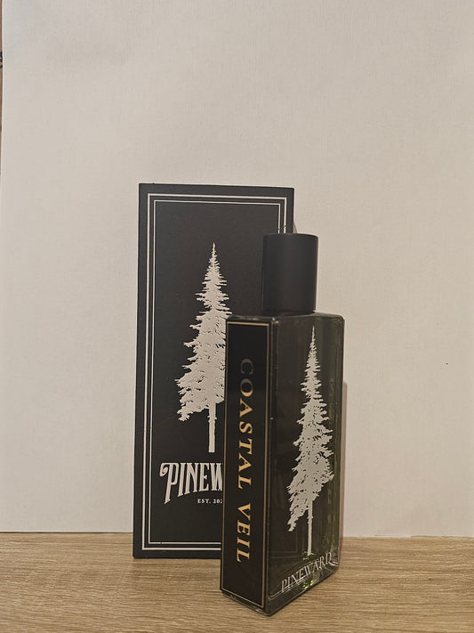 A bottle of Coastal Veil fragrance by Pinewind, standing in front of its matching black and white box on a light wood surface against a plain off-white wall. Pineward Coastal Veil| Fragrance Samples UK 1ml 2ml and 5ml perfume samples available 