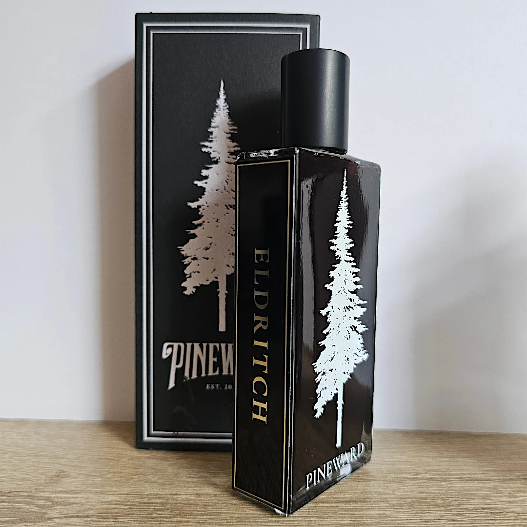 A bottle of Pinewood Elderitch fragrance stands in front of its matching black box on a light wood surface against a plain light gray wall. The packaging features a silver pine tree graphic and gold lettering.