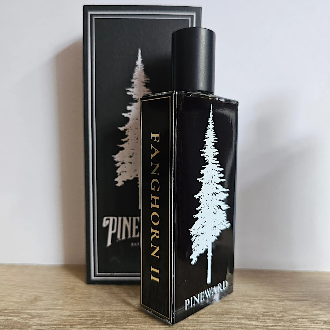 Fanghorn 2 Pinewood fragrance bottle and its matching black packaging box, both decorated with a white pine tree graphic and gold lettering, set on a wooden surface against a plain wall.