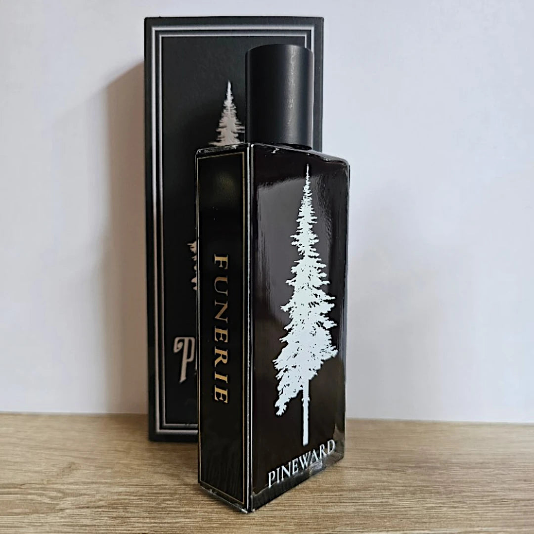 A black rectangular perfume bottle labeled 'FUNERIE' and 'PINWARD' with a white pine tree graphic, standing in front of its matching black box on a light wood surface against a plain light wall.