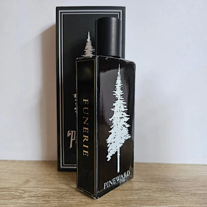 A black rectangular perfume bottle labeled 'FUNERIE' and 'PINWARD' with a white pine tree graphic, standing in front of its matching black box on a light wood surface against a plain light wall.