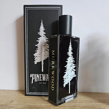 Pinewood Murkwood fragrance bottle and its matching black packaging box, both featuring a white pine tree graphic and gold lettering, set on a wooden surface against a plain wall.