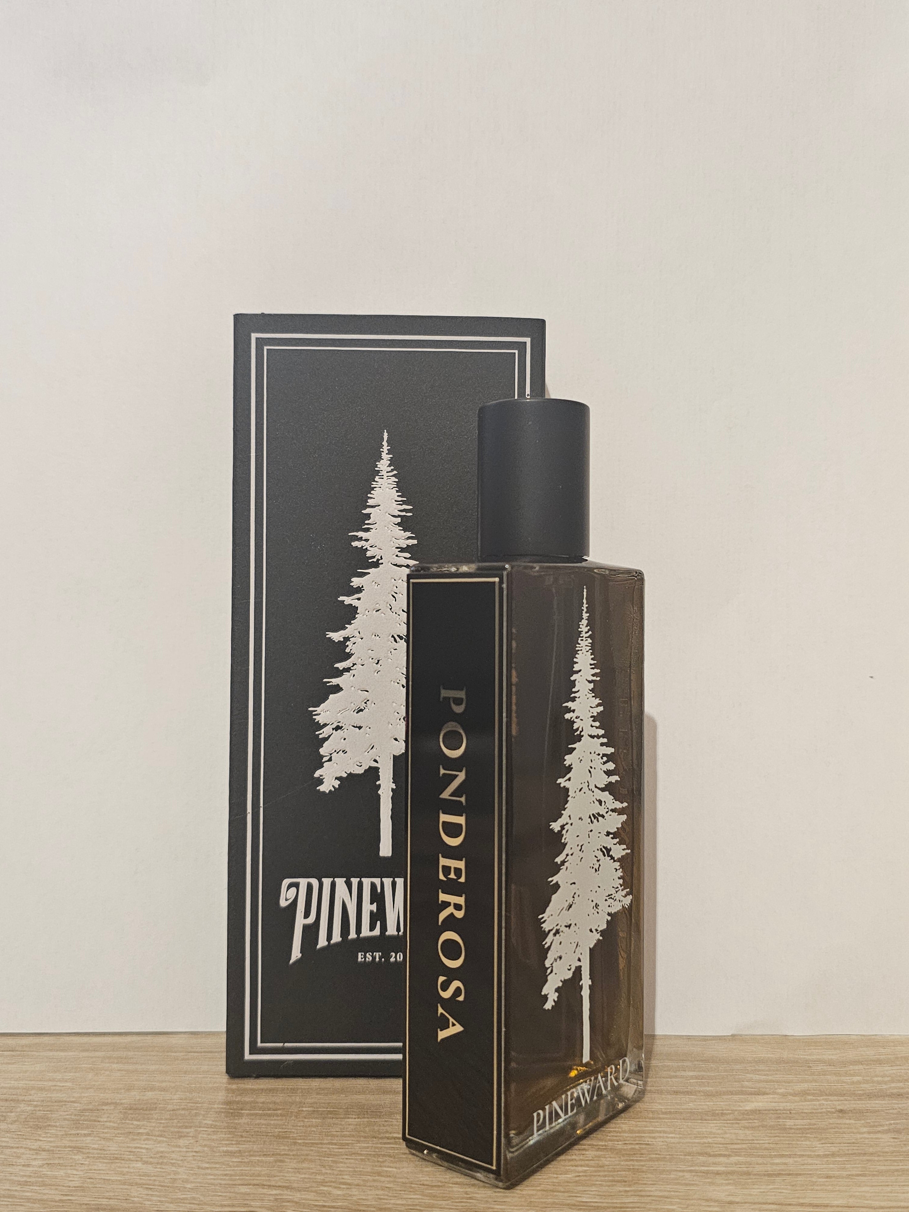 A bottle of Ponderosa fragrance oil or perfume next to its black packaging box, both featuring a white pine tree illustration and the brand name 'PINEWATER EST. 2014' on a light wood surface against a plain off-white wall. Pineward Ponderosa | Fragrance Samples UK 1ml 2ml and 5ml perfume samples available 