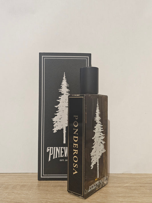 A bottle of Ponderosa fragrance oil or perfume next to its black packaging box, both featuring a white pine tree illustration and the brand name 'PINEWATER EST. 2014' on a light wood surface against a plain off-white wall. Pineward Ponderosa | Fragrance Samples UK 1ml 2ml and 5ml perfume samples available 