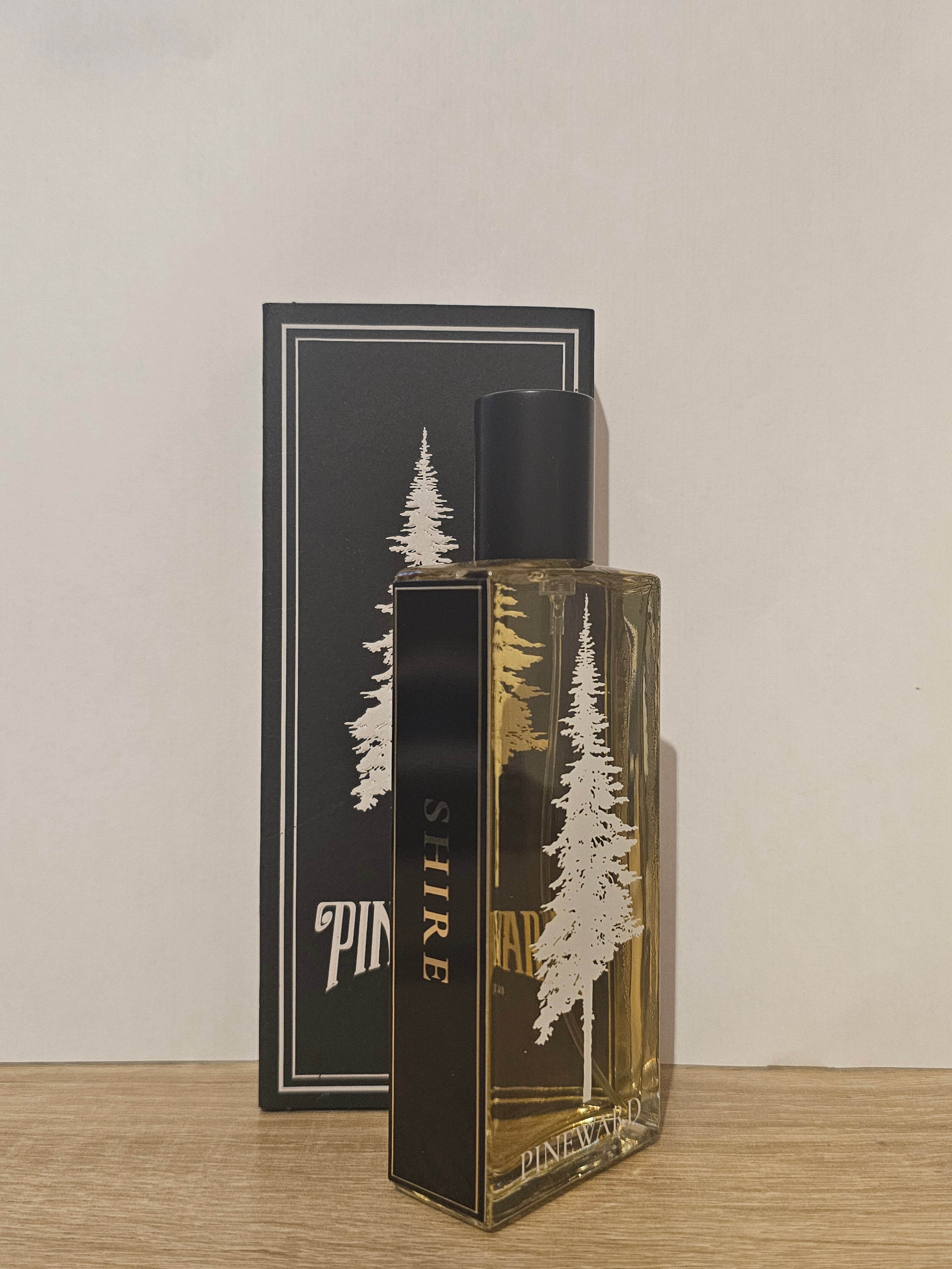 A rectangular glass perfume bottle with amber liquid and a black cap, standing in front of its black packaging box. The bottle and box feature a white pine tree illustration and the brand name 'SHIRE'. Pineward Shire | Fragrance Samples UK 1ml 2ml and 5ml perfume samples available 