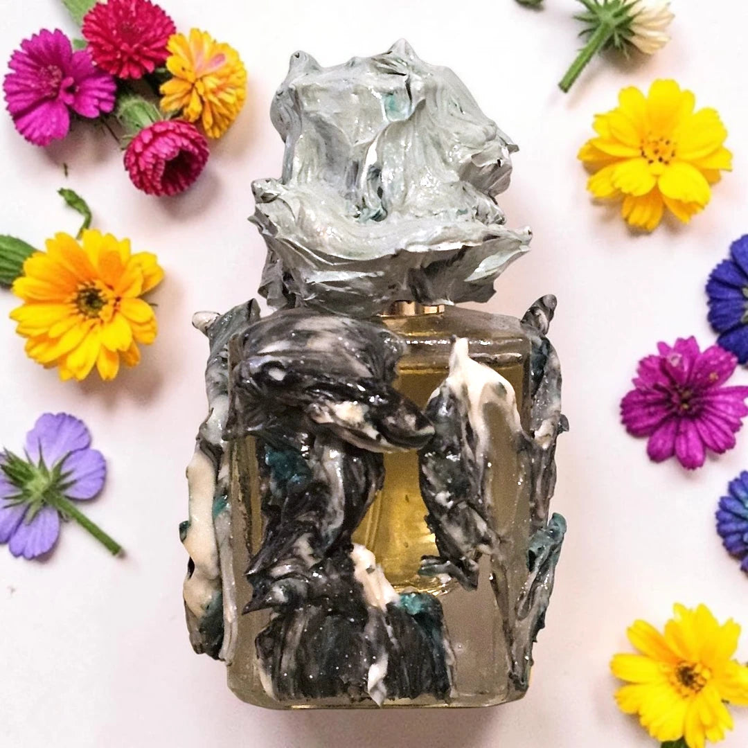 A sculptural perfume bottle with a textured, organic cap and body, featuring marbled black, white, and gray resin with hints of teal and gold. The bottle is surrounded by colorful artificial flowers on a light pink background. Solivagant Animal Farm | Fragrance Sample UK 1ml 2ml and 5ml perfume samples available 