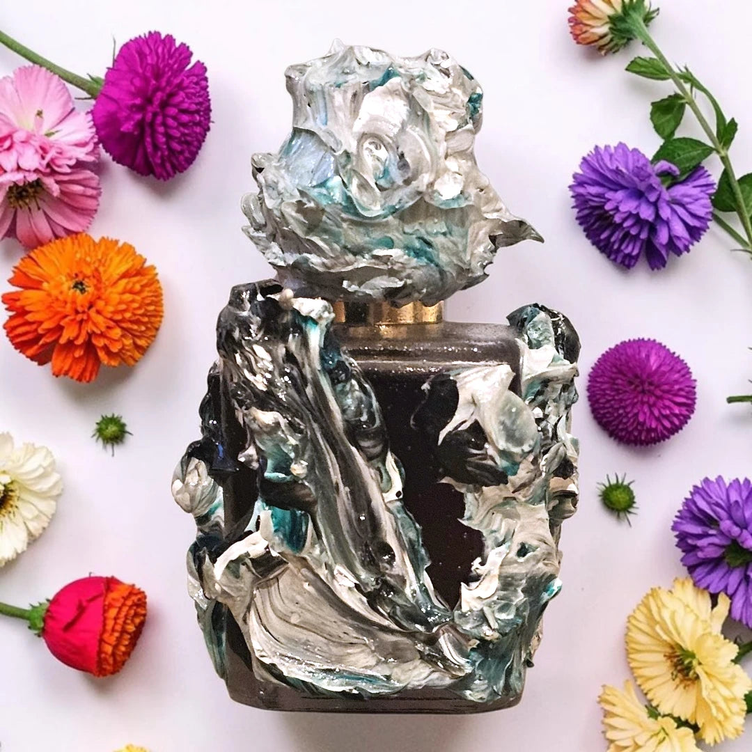 A sculptural perfume bottle with thick, textured impasto paint detailing in silver, black, and teal, surrounded by colorful artificial flowers on a white background. Solivagant Jason Voorhees | Fragrance Sample UK 1ml 2ml and 5ml perfume samples available 