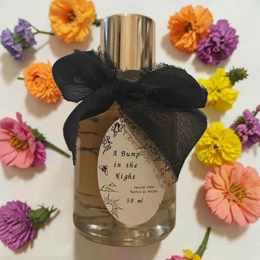A 50 ml bottle of 'A Bump in the Night' Eau de Parfum with a shiny gold cap, a large black sheer ribbon bow, and a cream-colored oval label. The bottle is centered and surrounded by a decorative flat-lay of colorful artificial flowers (orange, pink, yellow, purple) on a white background. Sorce A Bump In The Night | Fragrance Samples UK 1ml 2ml and 5ml perfume samples available 