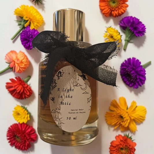 A 50ml bottle of 'A Light in the Attic' perfume by Parfum de Paris, featuring a gold cap, a black sheer ribbon bow, and a vintage-style cream label. The bottle is surrounded by colorful artificial flowers on a white background. Sorce A Light In The Attic | Fragrance Samples UK 1ml 2ml and 5ml perfume samples available 