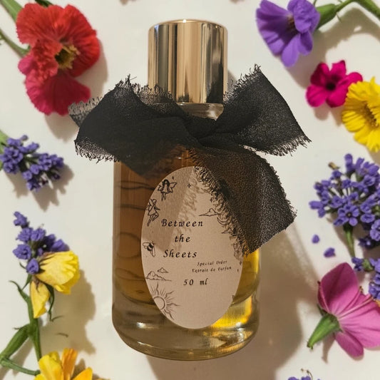 A 50 ml bottle of 'Between the Sheets' Eau de Parfum with a gold cap, a black tulle ribbon bow, and a beige label, surrounded by colorful artificial flowers on a white background. Sorce Between The Sheets | Fragrance Samples UK 1ml 2ml and 5ml perfume samples available 