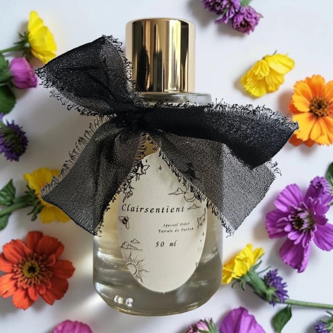 A 50 ml perfume bottle with a gold cap and a black sheer ribbon bow, labeled 'Clairsentient', styled on a white background with colorful flowers.| Fragrance Samples UK available in 1ml 2ml 5ml 10ml samples 