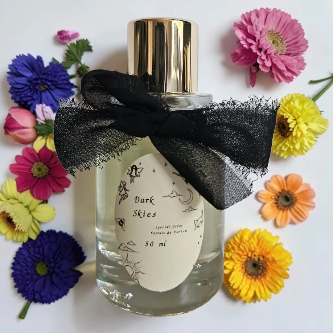 A 50 ml bottle of 'Dark Skies' special order perfume, featuring a clear glass body, a shiny gold cap, and a black sheer ribbon bow tied around the neck. The bottle is decorated with a cream-colored label and is surrounded by colorful artificial flowers on a white background.
 Fragrance Samples UK available in 1ml 2ml 5ml 10ml samples 
