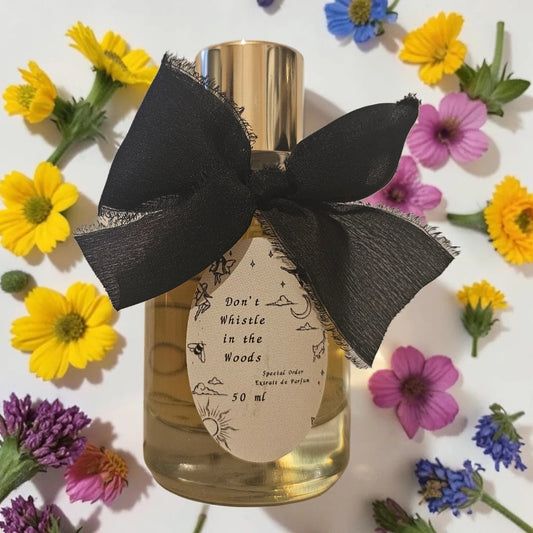 A 50ml bottle of 'Don't Whistle in the Woods' Eau de Parfum by Special Order, featuring a gold cap, clear glass showing golden-amber liquid, a beige paper label with black illustrations, and a large black sheer ribbon bow. The bottle is surrounded by yellow, pink, and purple flowers on a white background. Sorce Don't Whistle In The Woods | Fragrance Samples UK 1ml 2ml and 5ml perfume samples available 