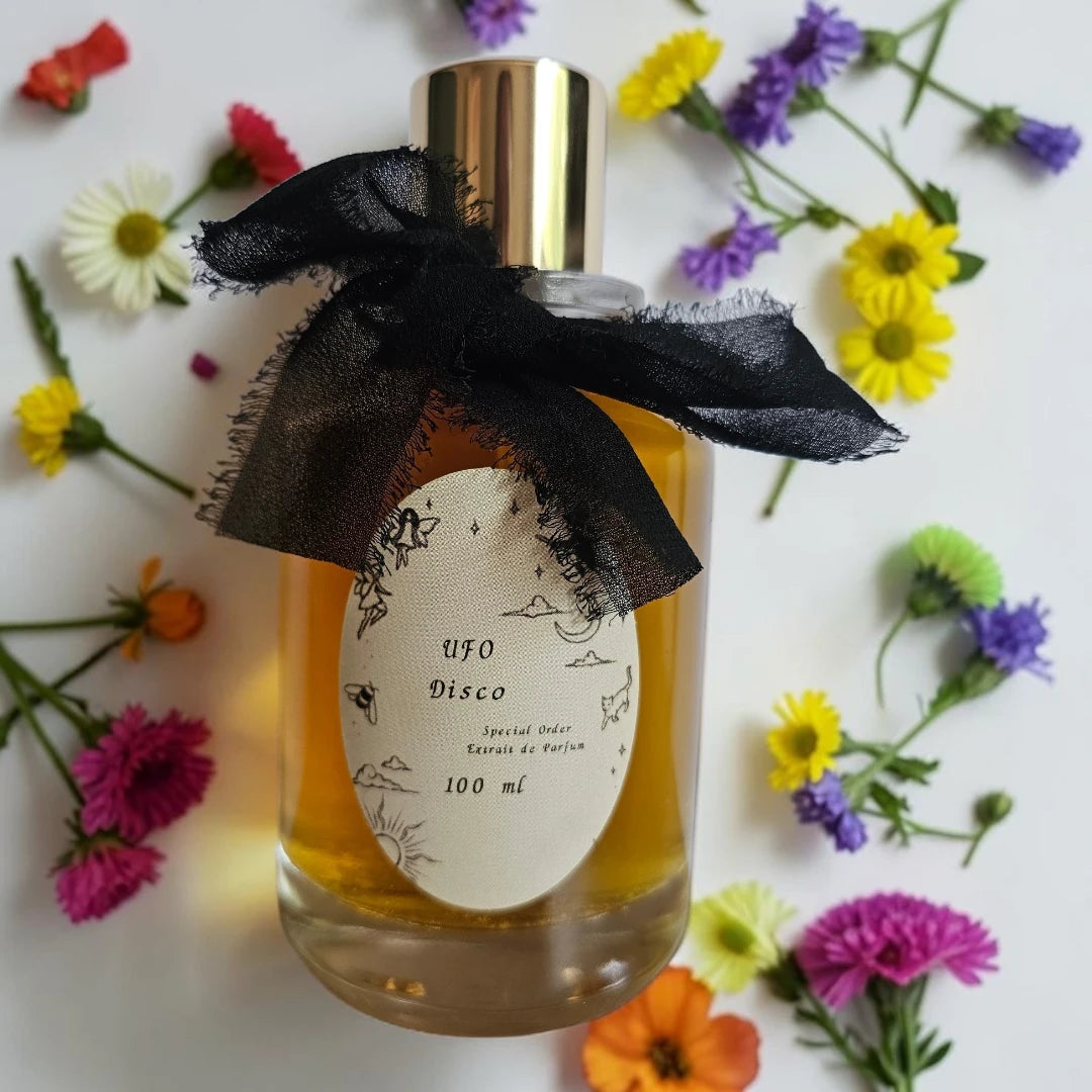 A 100 ml bottle of UFO Disco Eau de Parfum with a gold cap and a black sheer ribbon bow, surrounded by colorful artificial flowers on a white background. | Fragrance Samples UK available in 1ml 2ml 5ml 10ml samples 
