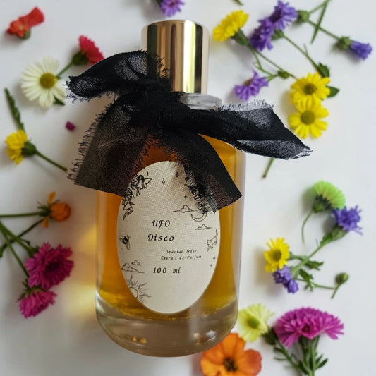 A 100 ml bottle of UFO Disco Eau de Parfum with a gold cap and a black sheer ribbon bow, surrounded by colorful artificial flowers on a white background. | Fragrance Samples UK available in 1ml 2ml 5ml 10ml samples 
