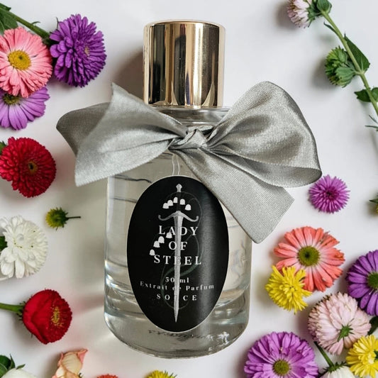 A 30ml bottle of Lady of Steel Extrait de Parfum with a shiny gold cap, a silver satin bow, and a black label, surrounded by colorful daisy flowers on a white background. Sorce lady of steel | Fragrance Samples UK 1ml 2ml and 5ml perfume samples available 