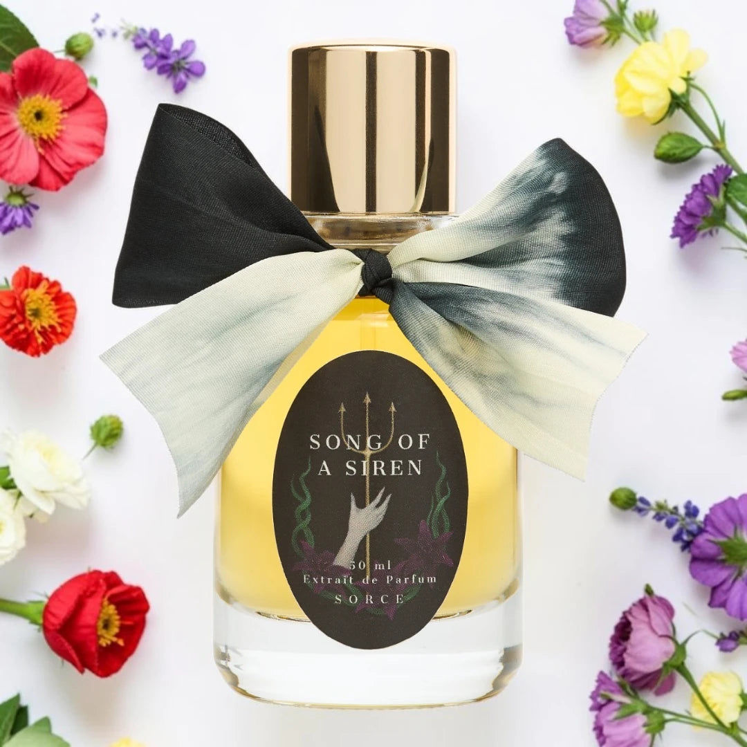 A bottle of Song of a Siren Extrait de Parfum with a gold cap and a large black and white tie-dye ribbon bow, surrounded by colorful flowers on a white background. Sorce song of a siren | Fragrance Samples UK 1ml 2ml and 5ml perfume samples available 